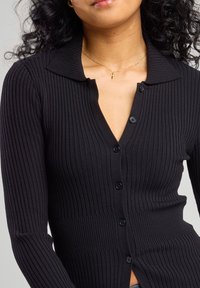 Black ribbed cardigan with a collar, featuring a V-neckline and six buttons. The fabric has a textured pattern, providing a snug fit.