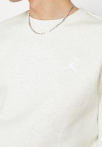 Närbild av en person som bär en ljusgrå sweatshirt med en liten vit Jumpman-logga på bröstet och en silverkedja.