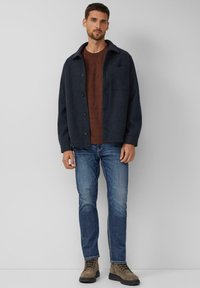 Dunkelblauer Wollmantel mit Knopfverschlüssen und einer Brusttasche, kombiniert mit einem rostbraunen Hemd und blauen Jeans, abgerundet mit braunen Schnürstiefeln.