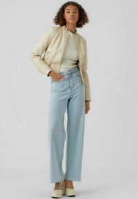 Giubbotto di pelle beige, top bianco a coste, jeans a gamba larga blu chiaro e scarpe con platform color crema. Capelli ricci, mani nelle tasche. Sfondo semplice.