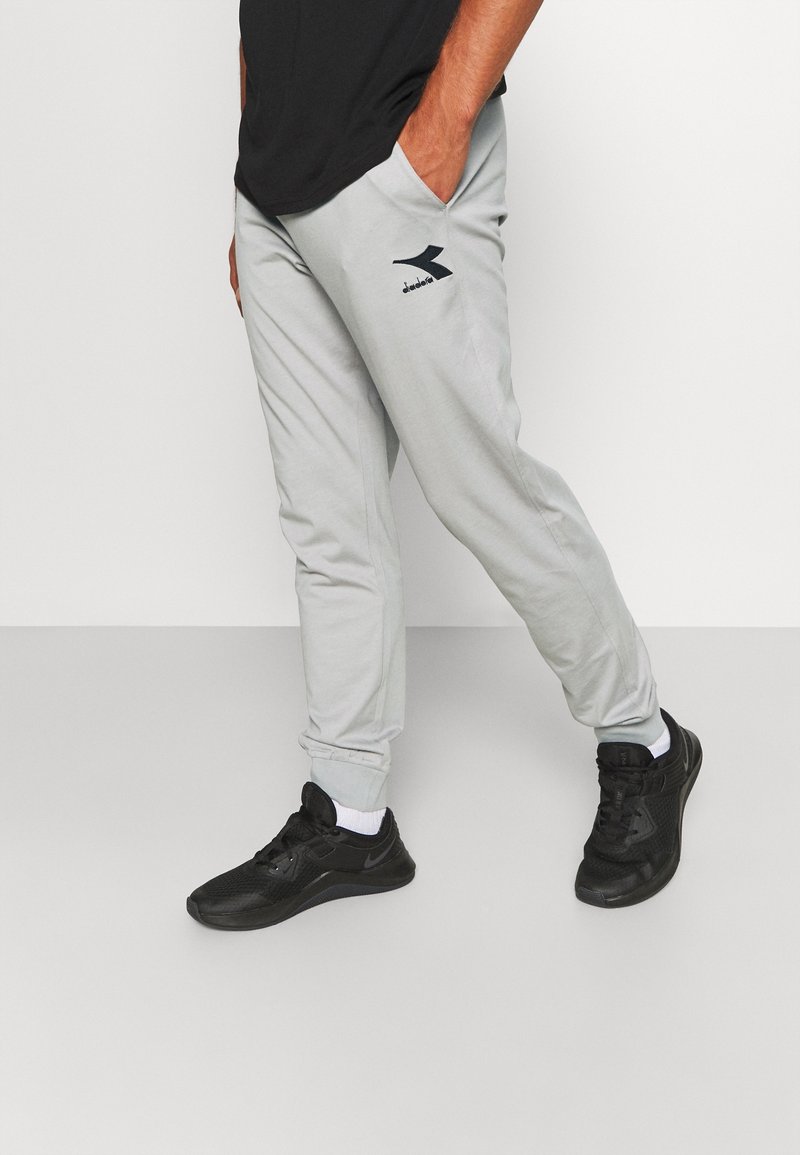 Man med ljusgrå joggingbyxor med en svart logotyp på låret, svarta sneakers med vita strumpor, stående mot en enkel bakgrund.