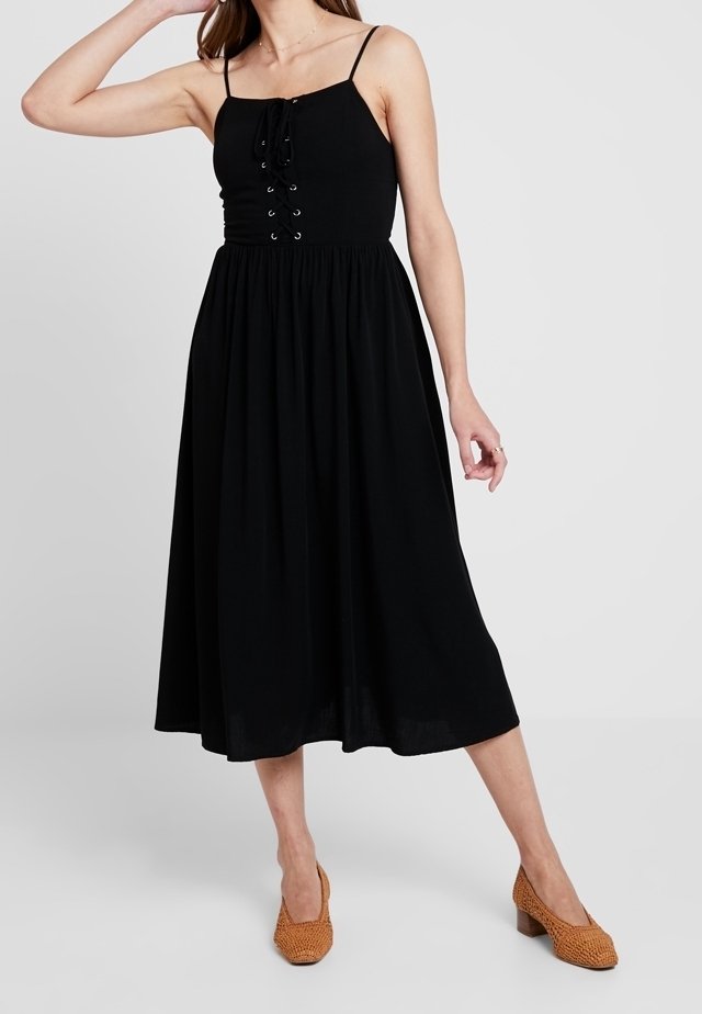 Femme portant une robe midi noire sans manches avec un détail à lacets sur le devant, associée à des chaussures basses tressées marron.