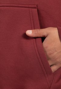 Tejido de sudadera burdeos con un bolsillo tipo canguro, que presenta costuras texturizadas y una mano descansando dentro del bolsillo.