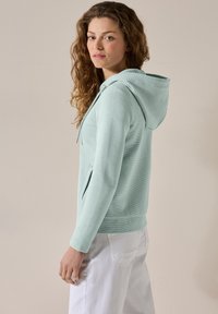 Felpa verde menta con tessuto texture, chiusura con zip e cappuccio regolabile; dotata di tasche laterali e polsini a coste, abbinata a pantaloni bianchi.