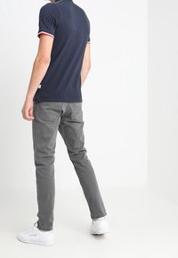 Polo shirt blu navy con rifiniture rosse e bianche, abbinato a jeans grigi. Il modello indossa sneakers bianche, mostrando un outfit casual di spalle.