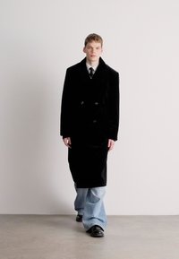 Διπλής σειράς μαύρο βελούδινο παλτό με μεγάλα κουμπιά, φορεμένο πάνω από άσπρη πουκάμισο και μαύρη γραβάτα, συνδυασμένο με ανοιχτόμπλε τζιν με φαρδιά πόδια και μαύρα παπούτσια.