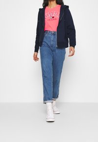 Felpa con zip navy sopra una maglietta rosa brillante "TOMMY JEANS", abbinata a jeans a vita alta di un blu chiaro e sneaker bianche.