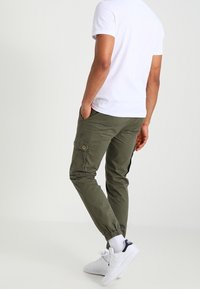 Kaki cargobyxor med elastiska muddar, med sidofickor och en liten cirkulär logotyp. Bärs med en enkel vit t-shirt och vita sneakers.