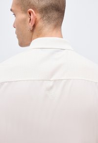 Άνδρας με κοντά μαλλιά και σκουλαρίκια, που κοιτάζει προς τα πίσω, φορώντας λευκό μπουφάν με διάτρητη πάνω πλάτη και ανάγλυφο λογότυπο αετού.