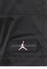 Svart tyg med en svart gummipatch med präglad "JORDAN" text och en rosa Jumpman-logotyp. Slät textur med synliga sömmar.