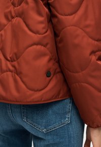 Κοντινή λήψη στην εσοχή και το μανίκι μπουφάν σε χρώμα σκουριάς με καπιτονέ ραφή, πάνω από τσεπάκι πίσω στα μπλε τζιν με ραμμένη σχεδίαση και ορατό χέρι.