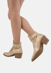 Botines metálicos dorados con tacón grueso, que presentan cortes, correas con hebillas y tachuelas decorativas. Textura suave y diseño de punta redonda.