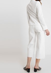 Bílá přizpůsobená bunda s vypasovaným střihem, spárovaná s bílými zkrácenými kalhotami. Černé sandály na podpatku mají zdobený pásek.