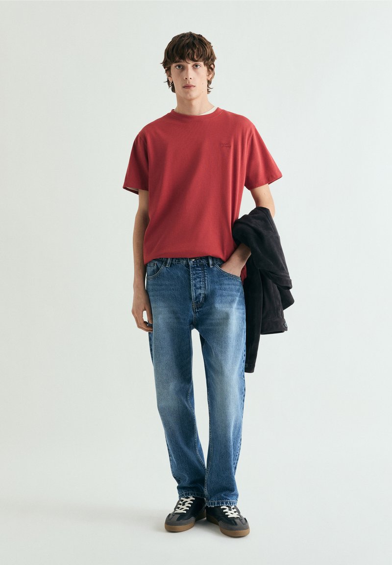 Jeune homme portant un t-shirt rouge, un jean bleu, des baskets noires, tenant une veste noire, debout devant un fond clair uni.