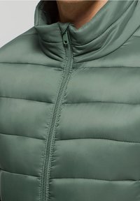 Prošívaná bunda v tlumené zelené barvě s vysokým límcem, vybavená centrálním zipem a hladkou, lesklou texturou.