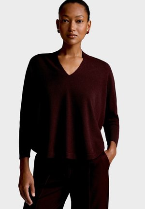 Femme portant un pull ample à col en V marron foncé à manches longues et un pantalon assorti sur un fond clair uni.