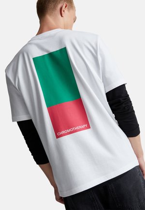 Een jongere draagt een wit T-shirt met groene en rode rechthoekige kleurvakken en het woord "CHROMOTHERAPY" op de achterkant.