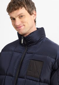 Giacca puffer blu navy con collo alto, chiusura a zip e patch logo nero testurizzato sul petto. Presenta un motivo a quadri cucito.