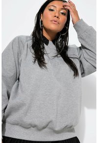 Felpa grigia oversized con una finitura testurizzata, dotata di scollo a girocollo e polsini a costine. Indossata sopra una camicia nera con colletto.
