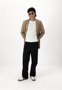 Blazer beige su un maglione bianco a coste, pantaloni neri e scarpe bianche con accenti grigi. Gli accessori includono occhiali da sole rotondi.