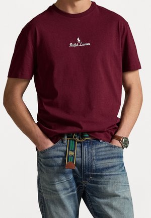 Homme portant un t-shirt bordeaux Ralph Lauren avec logo, jean bleu, ceinture verte rayée et une montre-bracelet avec bracelet en cuir marron.
