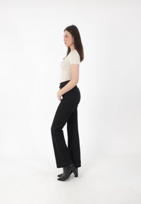 La modelo lleva una camisa ajustada beige de manga corta y unos pantalones vaqueros negros de pierna ancha y alta cintura, combinados con botines negros de tacón en una vista de perfil lateral.
