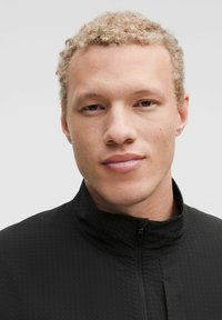 Černá zipová bunda s vysokým límcem, vyrobená z texturované látky. Model má světlé kudrnaté vlasy a neutrální výraz.