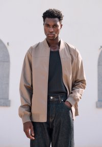 Standing man wearing a beige zip-up jacket over a black shirt and dark jeans, with one hand in his pocket, against a light background.