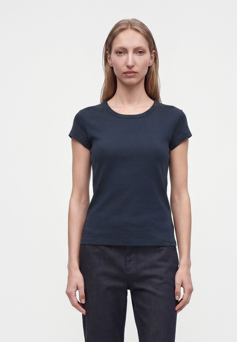 Femme aux longs cheveux blonds portant un t-shirt bleu marine uni à manches courtes et un pantalon foncé, debout devant un fond blanc.