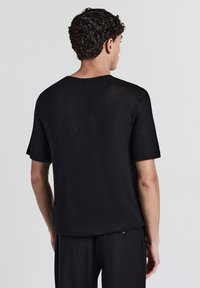 Uomo con capelli corti e ricci che indossa una camicia a maniche corte in maglia nera testurizzata e pantaloni neri, di spalle su uno sfondo bianco semplice.