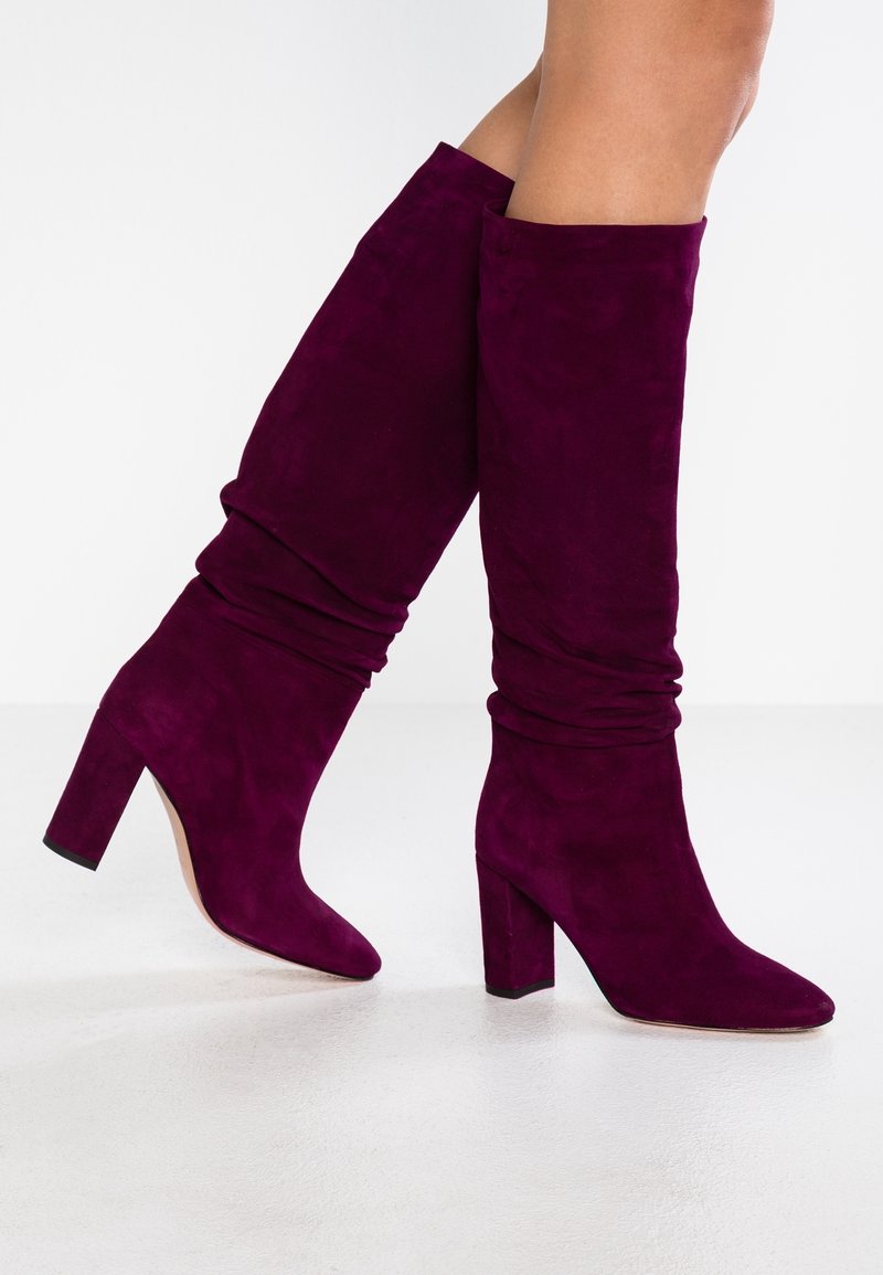 Botas de gamuza moradas hasta la rodilla con un diseño arrugado, punta afilada y tacón grueso, que presentan una textura suave y sin herrajes visibles.