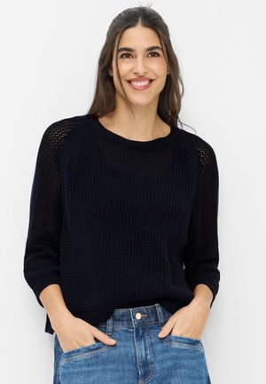 Smilende kvinde med langt mørkt hår iført en sort striktrøje og blå jeans, hænderne afslappet i lommerne, foran en ensfarvet hvid baggrund.