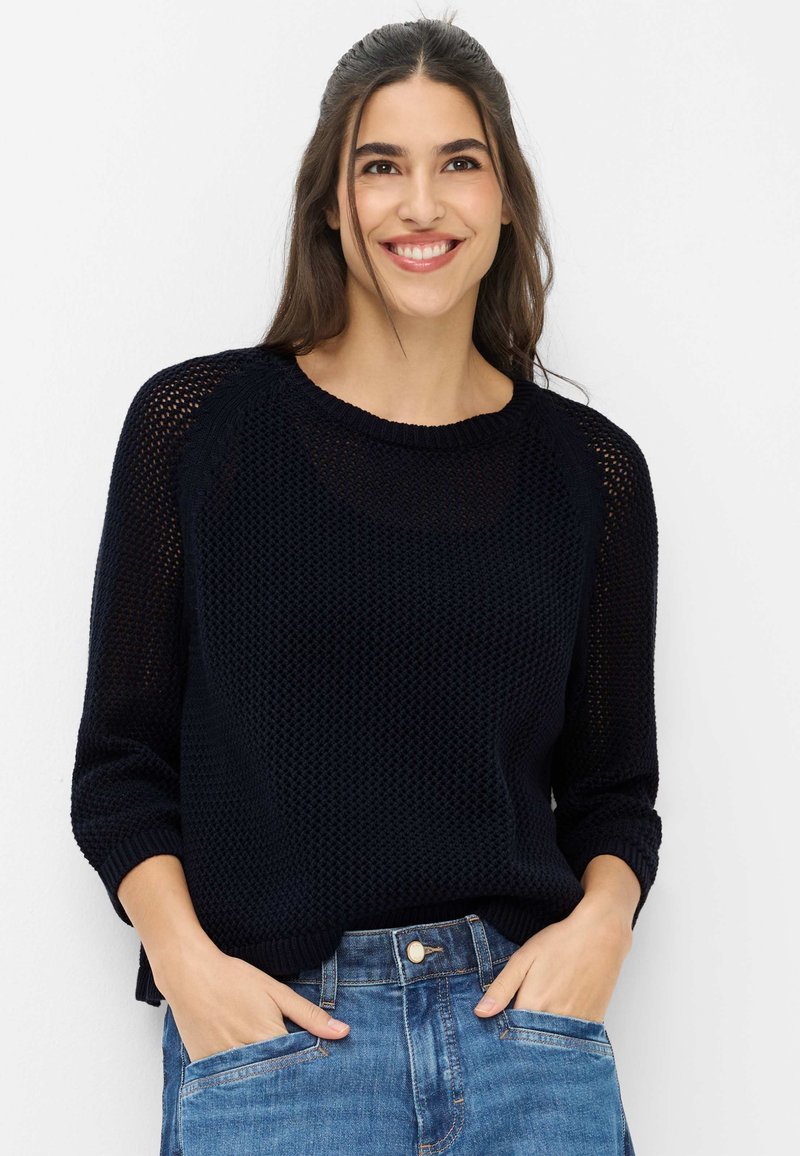 Femme souriante aux longs cheveux bruns, portant un pull en maille noir et un jean bleu, les mains décontractées dans les poches, sur un fond blanc uni.