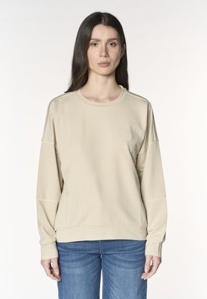 Donna con capelli lunghi e scuri che indossa una felpa beige e jeans blu, in piedi su uno sfondo bianco uniforme.