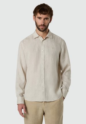 Hombre con cabello corto castaño y barba, vistiendo una camisa beige de manga larga abotonada y pantalones marrón claro, de pie frente a un fondo liso.