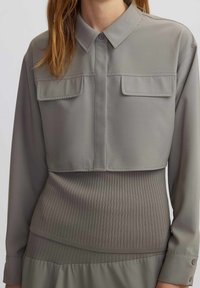 Femme portant une chemise boutonnée cropped taupe sur un haut en tricot côtelé à manches longues, se tenant devant un fond uni.