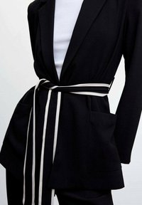 Blazer noir avec de grandes poches et une ceinture rayée noir et blanc nouée à la taille sur un top blanc côtelé.