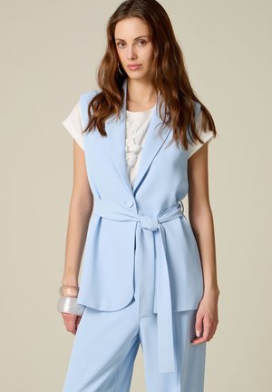 Femme aux longs cheveux bruns portant un blazer sans manches ceinturé bleu clair sur une chemise blanche à manches courtes, un pantalon bleu et des bracelets transparents.