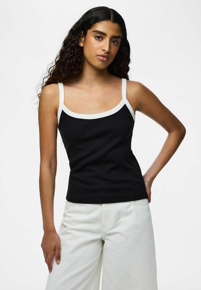 Femme aux longs cheveux bouclés portant un débardeur noir à bretelles blanches et un pantalon blanc, debout avec une main sur la hanche.