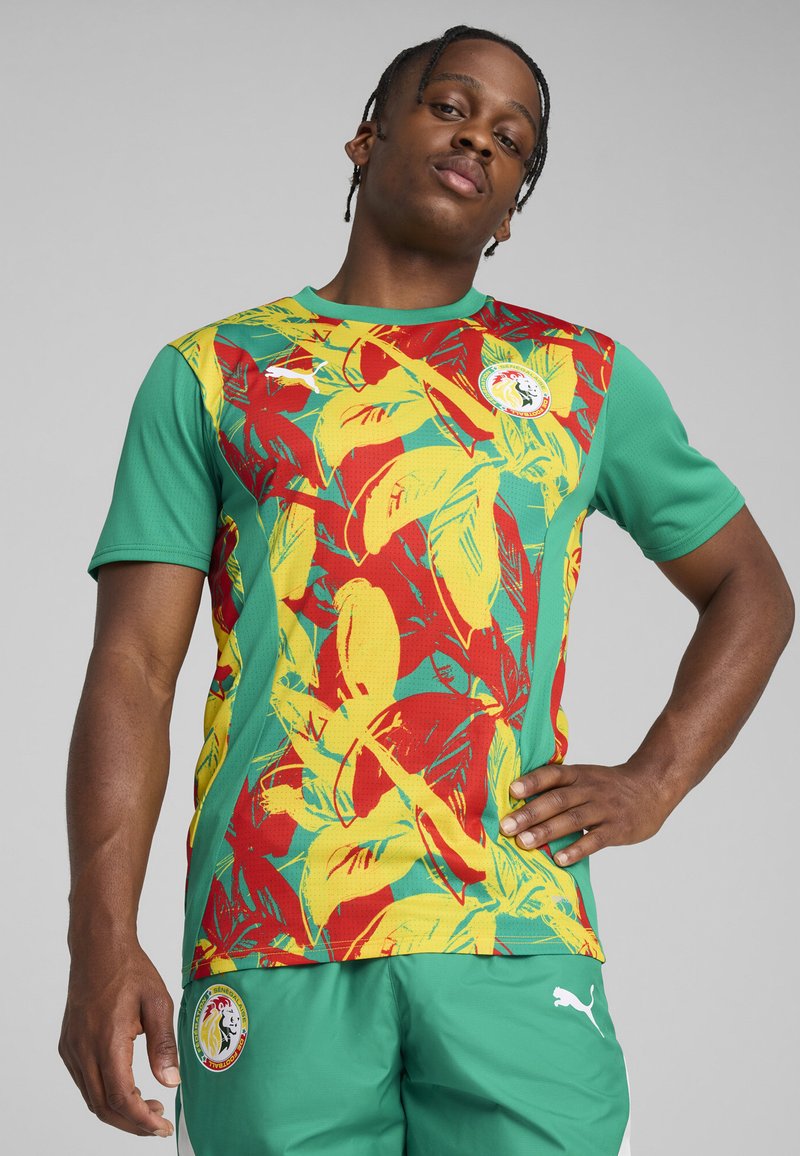 Jersey deportivo verde con vibrantes patrones de hojas amarillas y rojas, con un cuello redondo y un parche de logo en el pecho.
