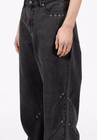 Czarne spodnie dżinsowe mają luźny krój, przednie kieszenie oraz srebrne oczka wzdłuż szwu, podkreślające charakterystyczny design.