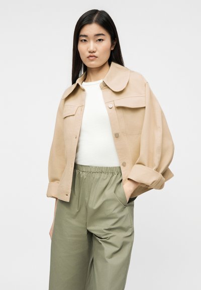 Νεαρή γυναίκα που φοράει μπεζ φαρδιά μπουφάν, λευκή μπλούζα και χακί παντελόνι, στέκεται με το ένα χέρι στην τσέπη μπροστά σε απλό φόντο.