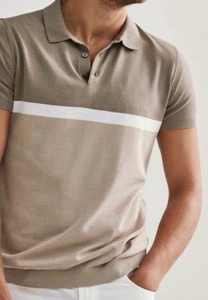 Homme portant un polo beige à manches courtes avec une rayure horizontale blanche, debout avec une main dans sa poche.