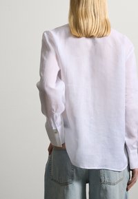 Camicia bianca di lino con maniche lunghe, caratterizzata da polsini con bottone e una vestibilità morbida, abbinata a pantaloni di denim azzurri.