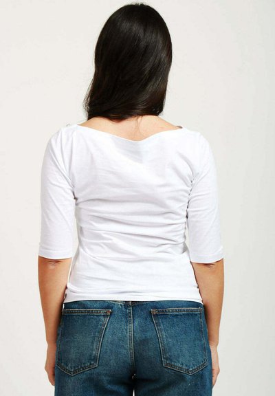 T-shirt bianco aderente con maniche a tre quarti e scollatura a barca. La vista posteriore mostra un tessuto liscio e cuciture sottili. Jeans in denim blu.