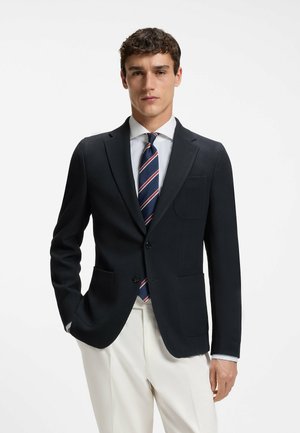 Unge mand iført en navy blazer, hvid skjorte, navy slip med striber og cremefarvede bukser, stående foran en ensfarvet hvid baggrund.