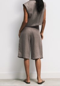 Mujer de pie frente a una pared blanca, vestida con un top sin mangas gris suelto, pantalones cortos a la altura de la rodilla a juego y sandalias negras de tacón abierto.