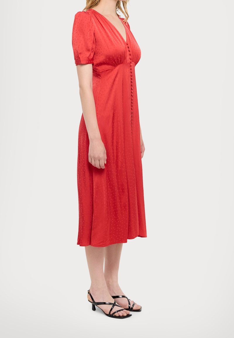 Mujer vestida con un vestido midi rojo con botones en la parte delantera y mangas cortas abullonadas, combinado con sandalias negras de tacón bajo y tiras.