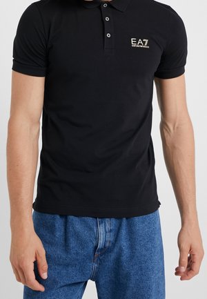 Homme portant un polo noir EA7 Emporio Armani et un jean bleu, debout avec les bras détendus le long du corps.