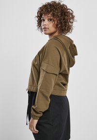 Olíva zöld, rövidített kapucnis pulóver, húzózsinórral az alján és széles ujjal, puha tapintással. Sötét szoknyával párosítva.